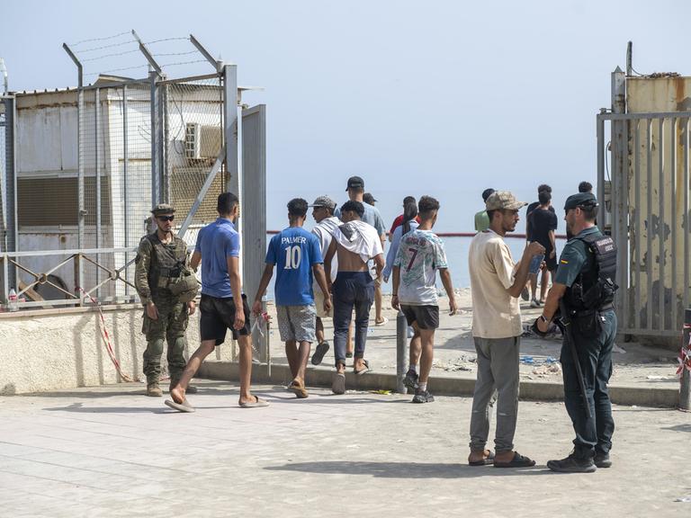 Mehrere Männer gehen durch einen abgesperrten Kontrollbereich in Ceuta. Spanische Sicherheitskräfte stehen neben ihnen. Im Hintergrund sind hohe Metallzäune und das Meer zu sehen. Mehrere Männer gehen durch einen abgesperrten Kontrollbereich in Ceuta. Spanische Sicherheitskräfte stehen neben ihnen. Im Hintergrund sind hohe Metallzäune und das Meer zu sehen.
