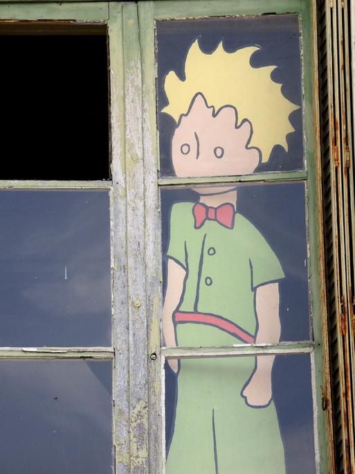 Die Figur des "Kleinen Prinzen" mit gelben Haaren und grüner Kleidung blickt hinter einem Fenster nach draußen.