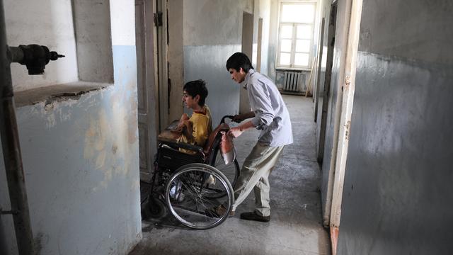 Ein Jugendlicher fährt einen Jungen im Roll-Stuhl von einem Flur in ein Zimmer. Das Gebäude ist in keinem guten Zustand.
