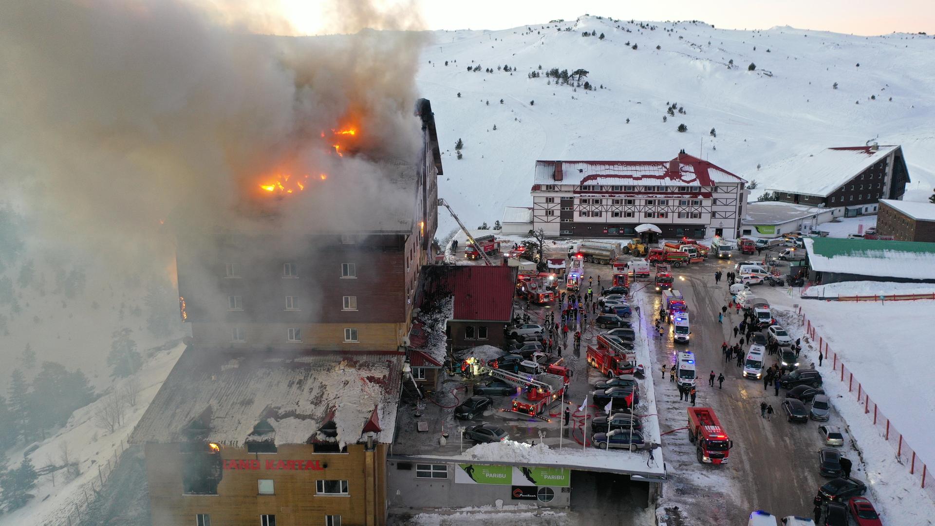 Blick von oben auf das brennende Hotel im türkischen Ski Resort Kartalkaya. Auf der Straße stehen viele Feuerwehr- und Polizeifahrzeuge, im Hintergrund Berge mit Schnee bedeckt.