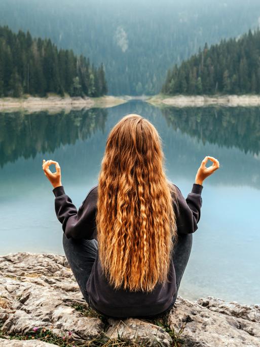 Eine Frau meditiert auf Steinen, am Ufer eines Waldumsäumten Sees, vielleicht sogar in den Bergen. Eine Frau meditiert auf Steinen, am Ufer eines Waldumsäumten Sees, vielleicht sogar in den Bergen.