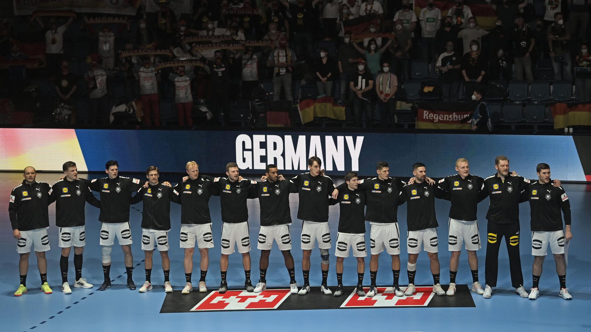 Die Spieler in schwarzen Trainingsjacken stehen nebeneinander und Arm in Arm auf dem Platz. Hinter ihnen der Schriftzug "Germany" und Zuschauer im Dunkeln auf den Tribünen. Manche davon mit Deutschland-Fahnen. 