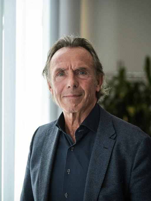 Thomas Druyen trägt ein dunkles Hemd, ein dunkles Sakko und steht vor einem mit Gardinen verhangenen Fenster.