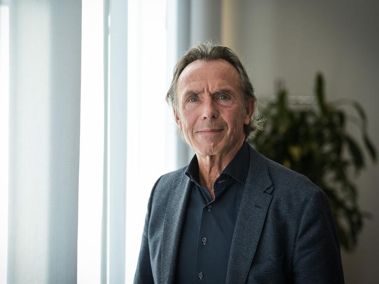Thomas Druyen trägt ein dunkles Hemd, ein dunkles Sakko und steht vor einem mit Gardinen verhangenen Fenster.