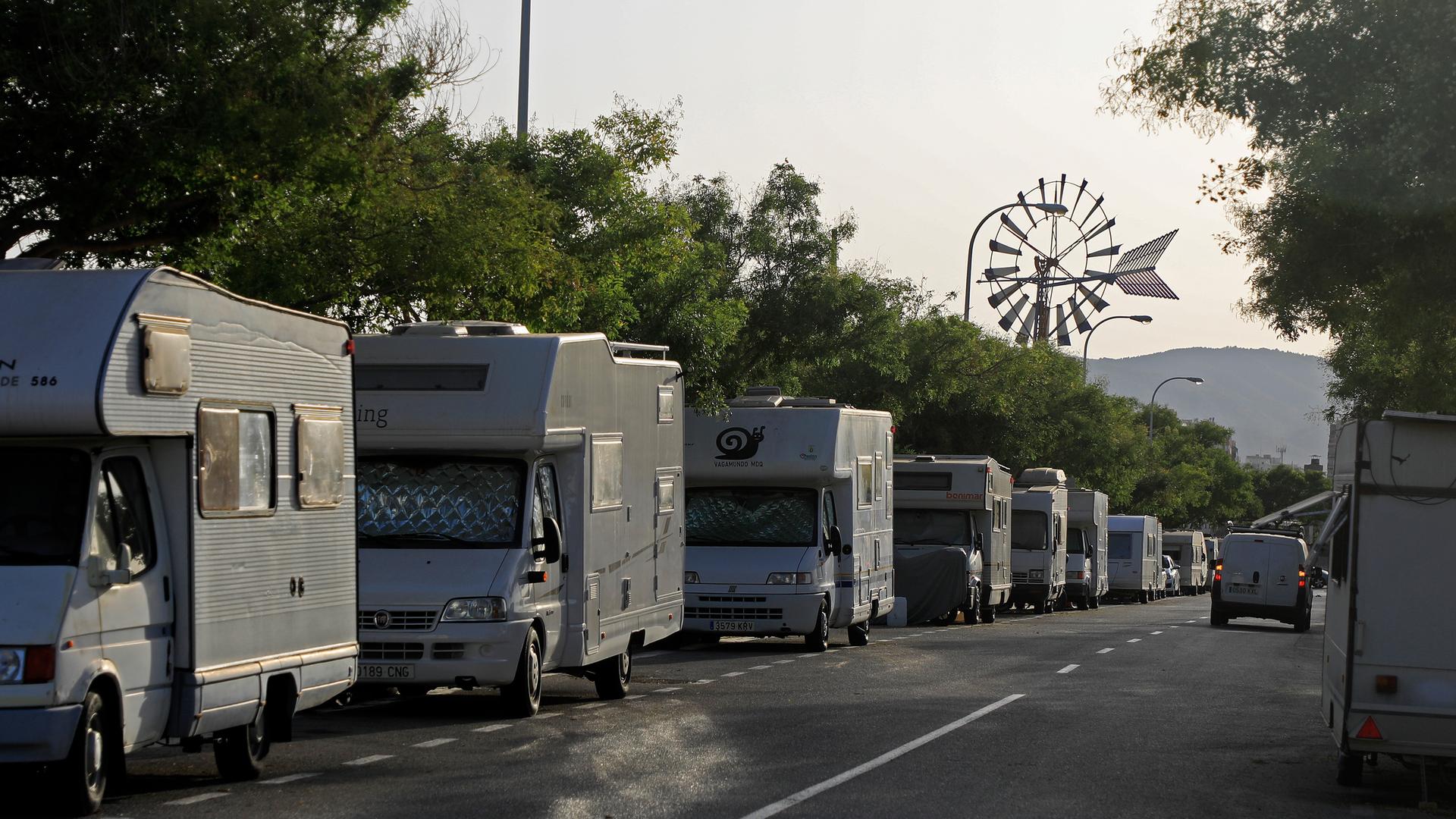 Blick in eine von Wohnmobilen gesäumte Straße. Am Ende der Straße ist zwischen den Bäumen ein Windrad zu sehen.