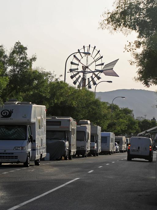 Blick in eine von Wohnmobilen gesäumte Straße. Am Ende der Straße ist zwischen den Bäumen ein Windrad zu sehen.