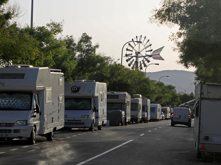 Blick in eine von Wohnmobilen gesäumte Straße. Am Ende der Straße ist zwischen den Bäumen ein Windrad zu sehen.