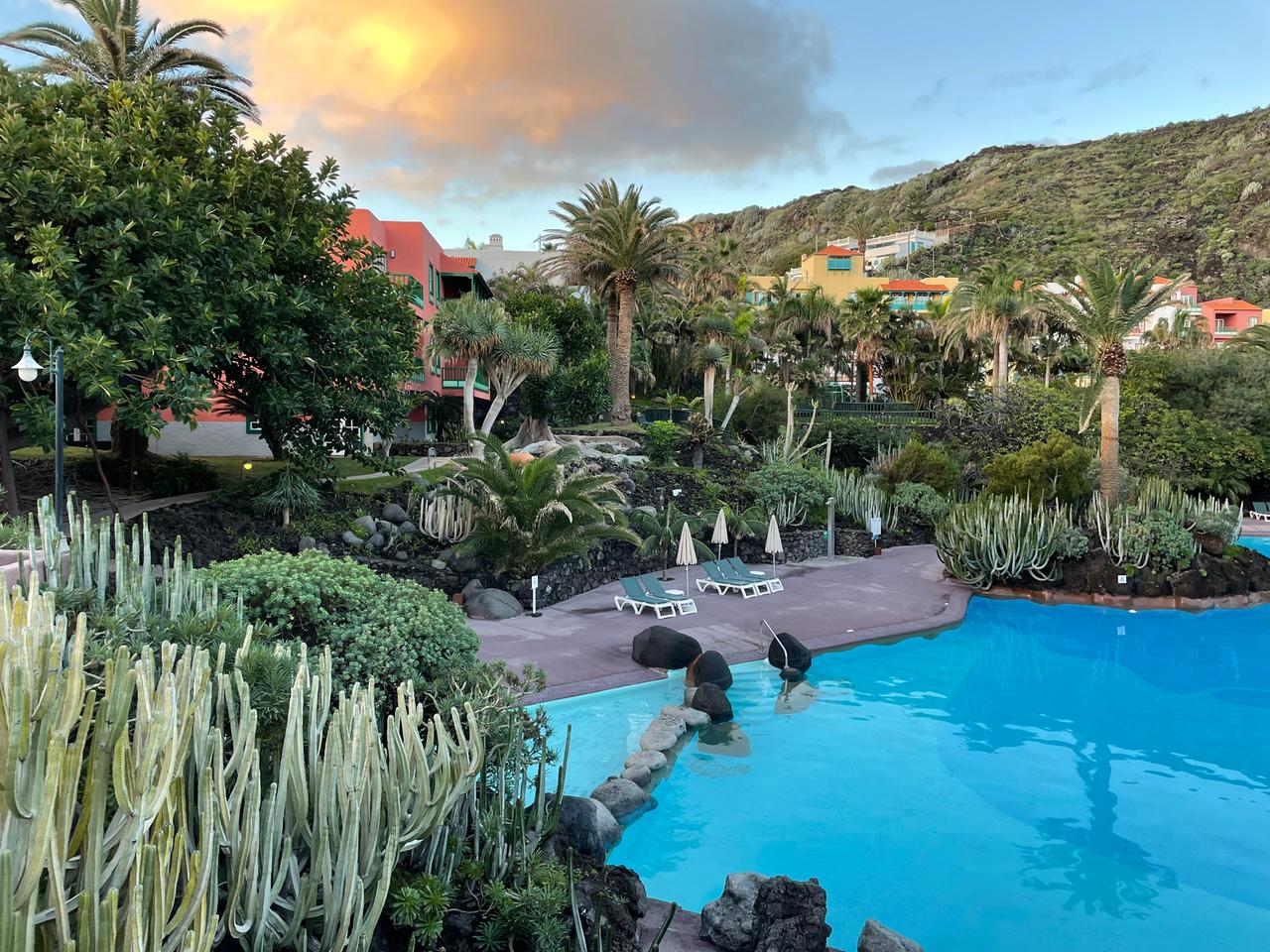 Ein idyllischer Hotelpool mit Palmen und Liegen drumherum. Im Hintergrund der vom Vulkan rotgefärbte Himmel.