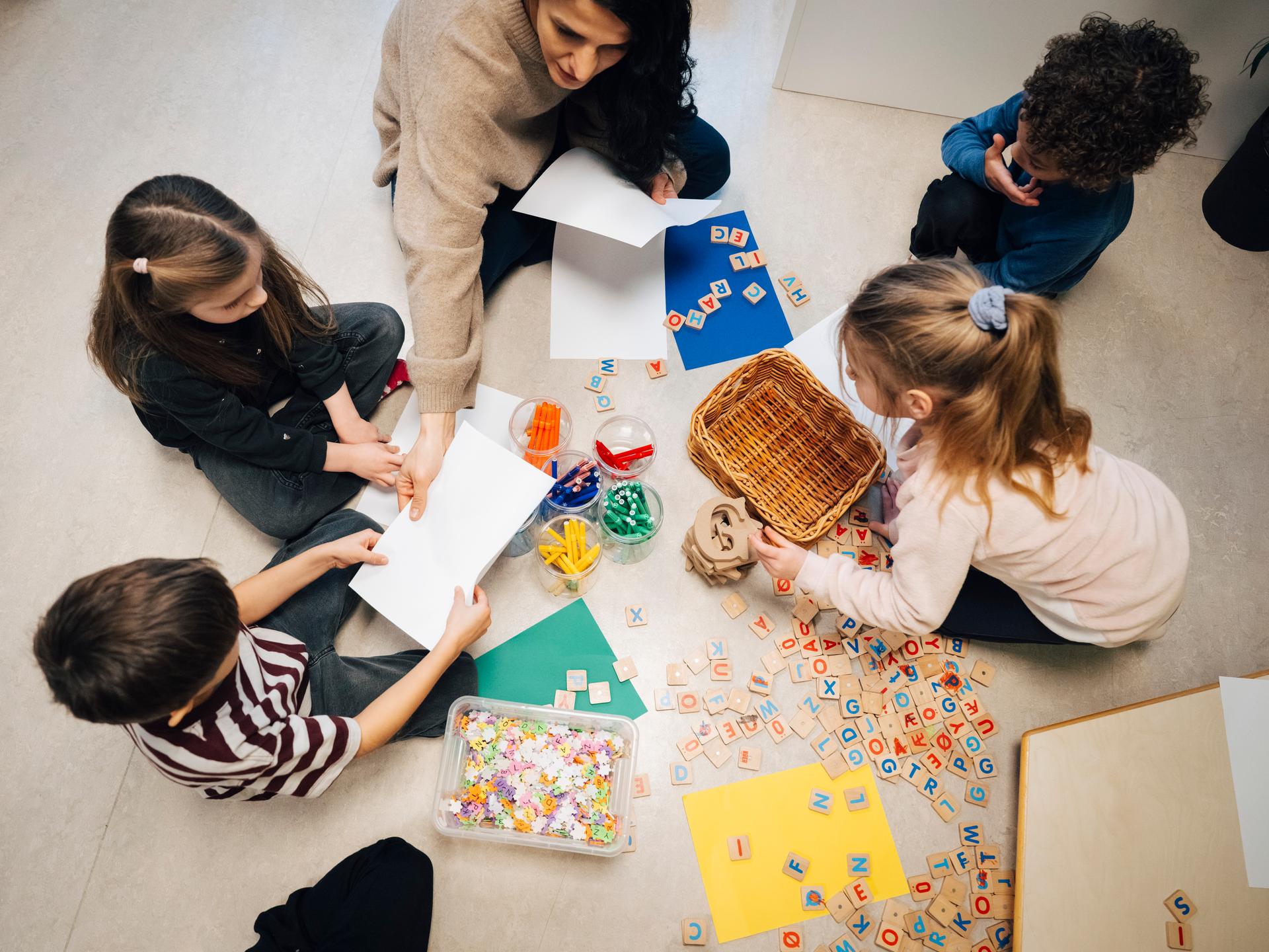 Eine Lehrerin sitzt in einem Klassenzimmer mit einigen Kindern am Boden. Sie arbeiten mit Holzplättchen, auf die Buchstaben gedruckt sind.