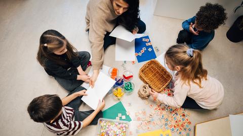 Eine Lehrerin sitzt in einem Klassenzimmer mit einigen Kindern am Boden. Sie arbeiten mit Holzplättchen, auf die Buchstaben gedruckt sind.
