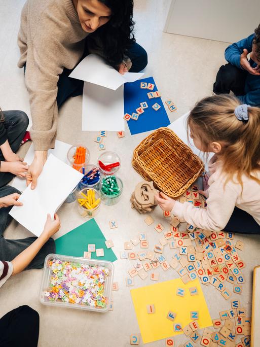 Eine Lehrerin sitzt in einem Klassenzimmer mit einigen Kindern am Boden. Sie arbeiten mit Holzplättchen, auf die Buchstaben gedruckt sind.
