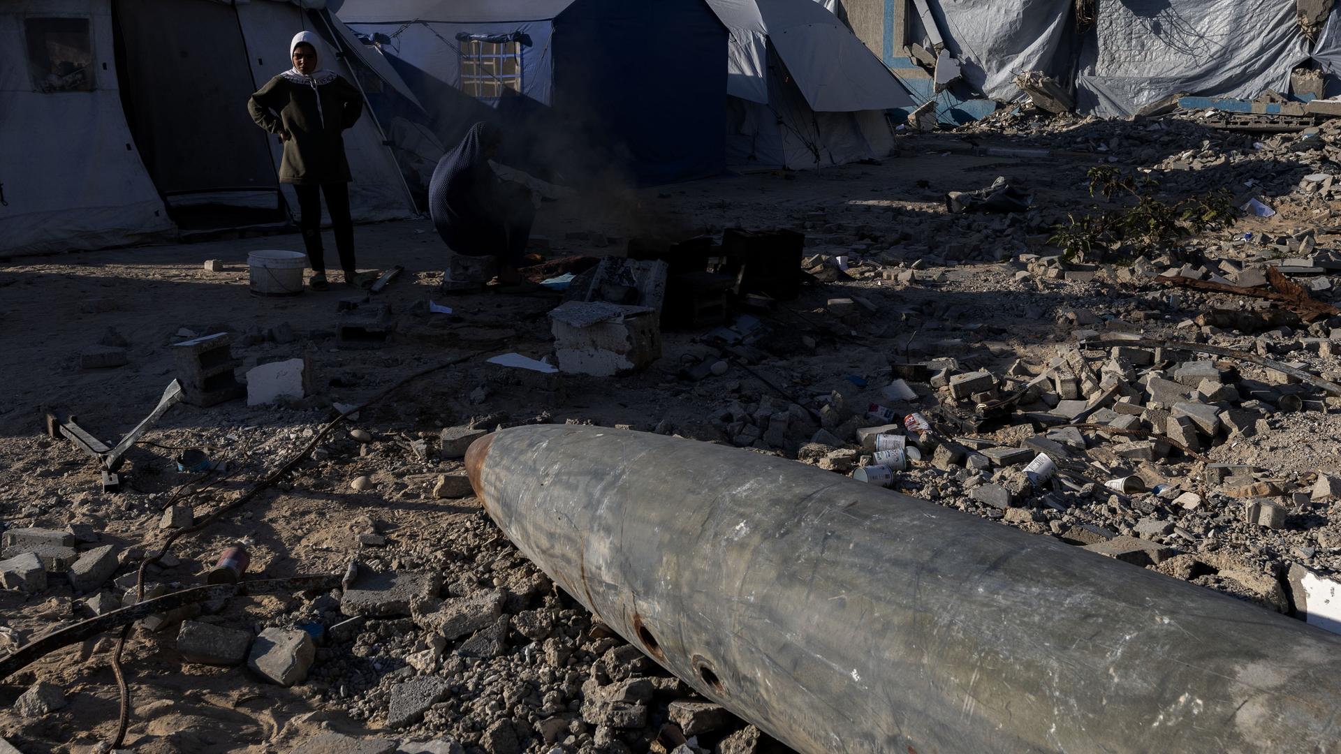 Im Vordergrund liegt eine Bombe, die von der israelischen Armee zurückgelassen wurde und nicht explodiert ist. Sie zeigt im Hintergrund auf Flüchtlingszelte im Gazastreife. Davor steht eine Person.