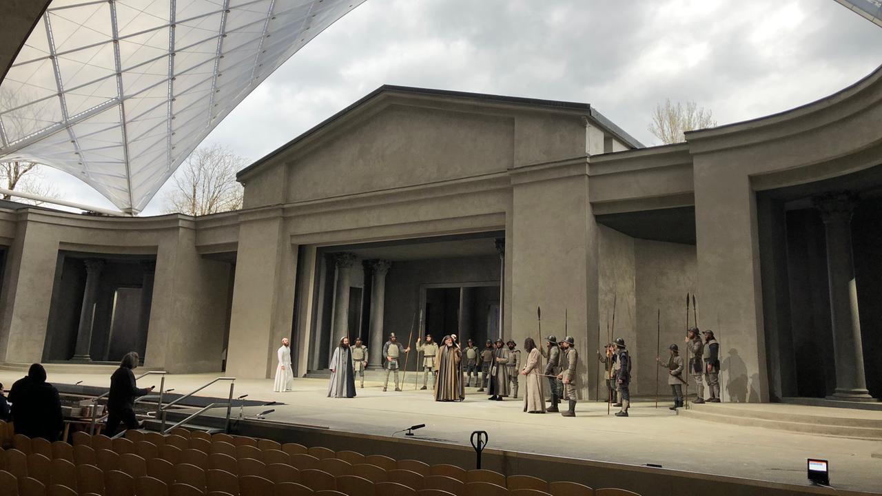 Auf der Bühne in Oberammergau stehen Dutzende Schauspieler. Das Dach für das Publikum wölbt sich links im Bild, über der Bühne ist der Himmel zu sehen, ganz im Vordergrund Stühle.