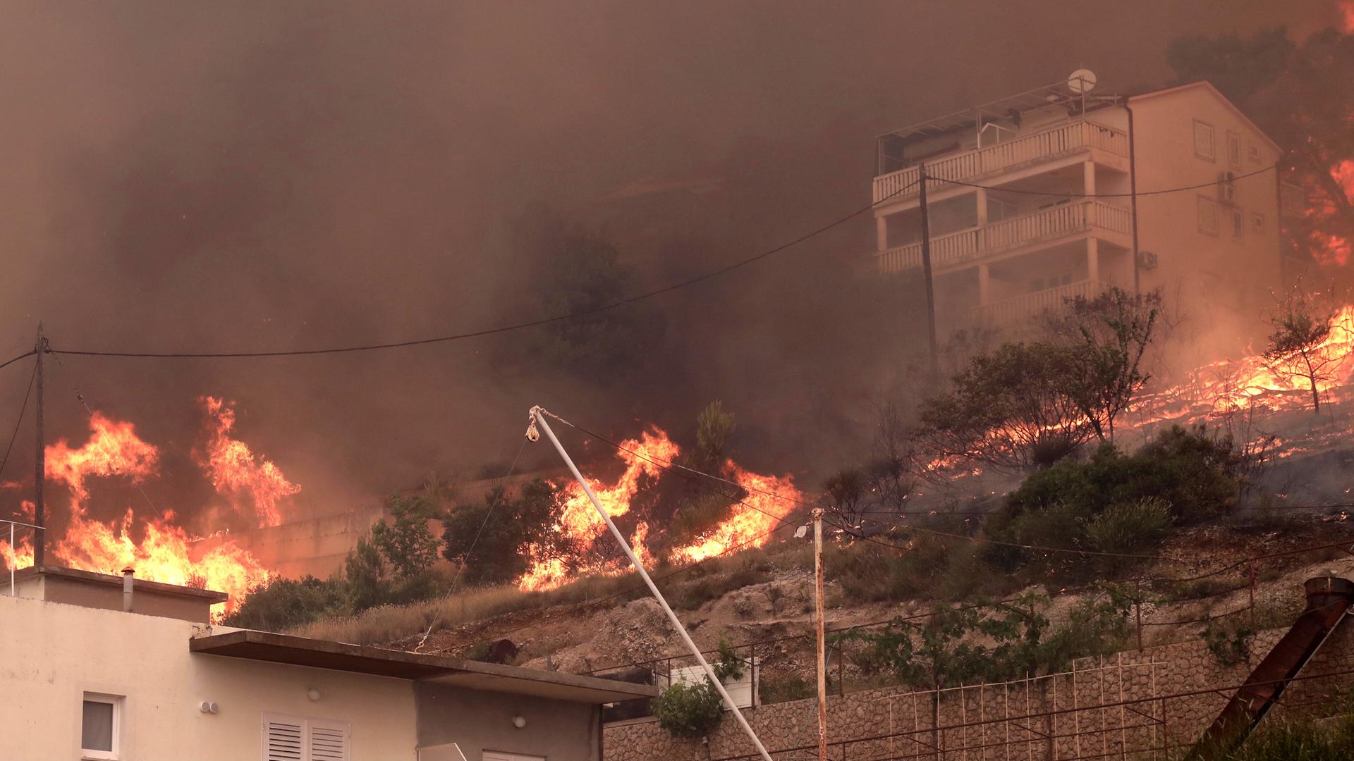 Feuer lodert zwischen Gebäuden. Ein heftiger Vegetationsbrand wütet an der kroatischen Adria-Küste und führt dazu, dass viele Touristen fliehen. Feuer lodert zwischen Gebäuden. Ein heftiger Vegetationsbrand wütet an der kroatischen Adria-Küste und führt dazu, dass viele Touristen fliehen.