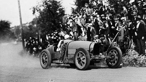 Diese Schwarz-Weiß-Aufnahme stammt aus dem Jahr 1927 und zeigt einen Rennwagen, der um eine Kurve fährt. Zuschauer stehen am Rand. Der Wagen verfügt über eine stromlinienförmige Karosserie.