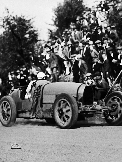 Diese Schwarz-Weiß-Aufnahme stammt aus dem Jahr 1927 und zeigt einen Rennwagen, der um eine Kurve fährt. Zuschauer stehen am Rand. Der Wagen verfügt über eine stromlinienförmige Karosserie.