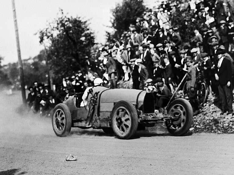 Diese Schwarz-Weiß-Aufnahme stammt aus dem Jahr 1927 und zeigt einen Rennwagen, der um eine Kurve fährt. Zuschauer stehen am Rand. Der Wagen verfügt über eine stromlinienförmige Karosserie.