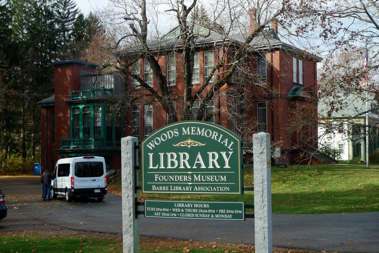 Blick auf ein Backsteingebäude vor dem auf einem grünen Schild "Woods Memorial Library" steht.