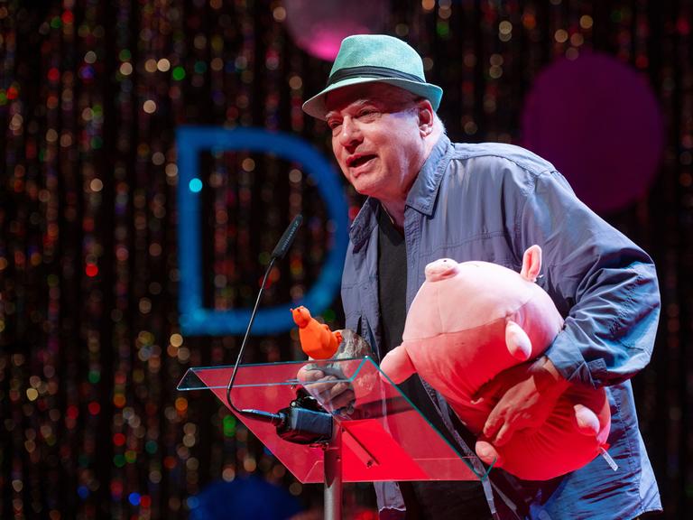 Rosa von Praunheim spricht mit Hut auf dem Kopf bei der Verleihung der 39. Teddy Awards am Rande der Berlinale und hält dabei ein rosa Stoffschwein im Arm.