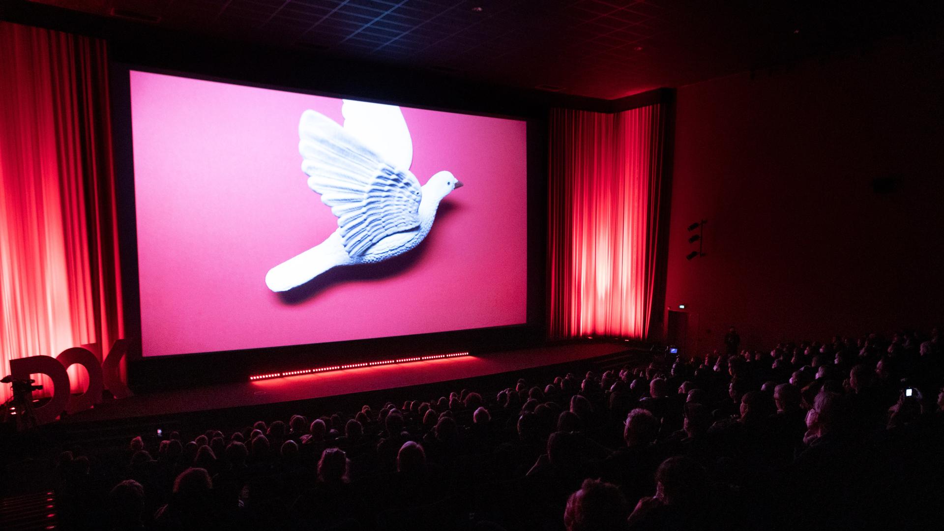 Eröffnungszeremonie des Filmfestivals DOK Leipzig. Zu sehen ist ein voller Kinosaal mit einer großen Taube auf der Leinwand.