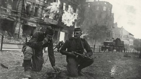 Soldaten der Roten Armee knien vor einem brennenden Haus in Berlin. Einer hält einen Hörer ans Ohr, der andere hat ein Maschinengewehr in der Hand.  