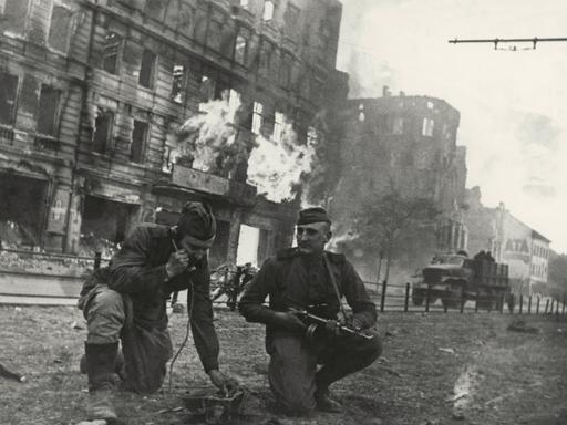 Soldaten der Roten Armee knien vor einem brennenden Haus in Berlin. Einer hält einen Hörer ans Ohr, der andere hat ein Maschinengewehr in der Hand.  