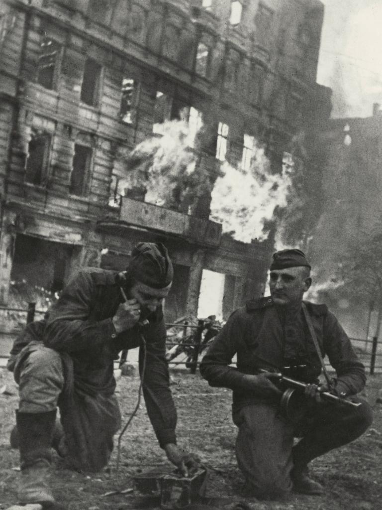 Soldaten der Roten Armee knien vor einem brennenden Haus in Berlin. Einer hält einen Hörer ans Ohr, der andere hat ein Maschinengewehr in der Hand.  
