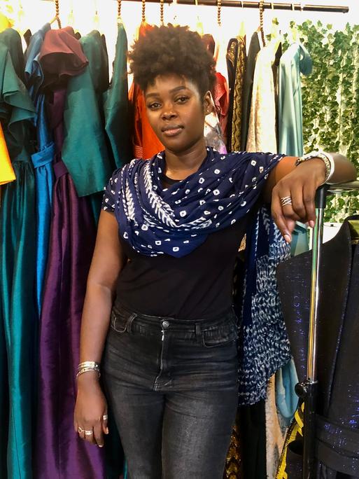 Eine schwarze Frau steht vor einer Kleiderstange, an der bunte Kleider hängen.