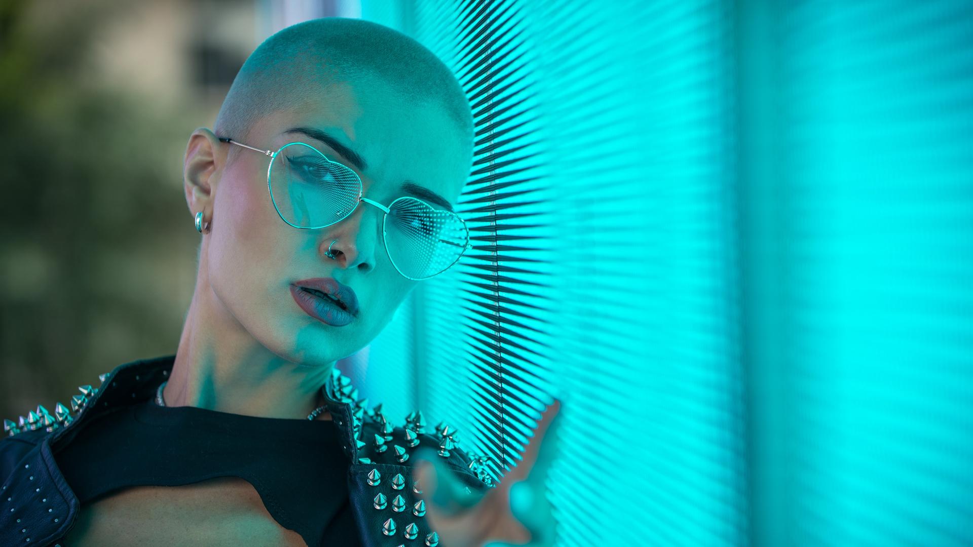 Portrait einer jungen Frau mit kahl geschorenem Kopf, Nietenjacke und Herzchen-Brille vor einem blaustichigen Hintergrund.