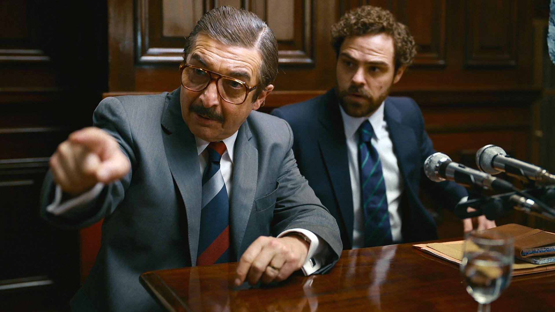 Zwei Männer in Anzügen in einem Gerichtssaal blicken auf ein Gegenüber. Der linke zeigt mit dem Finger.