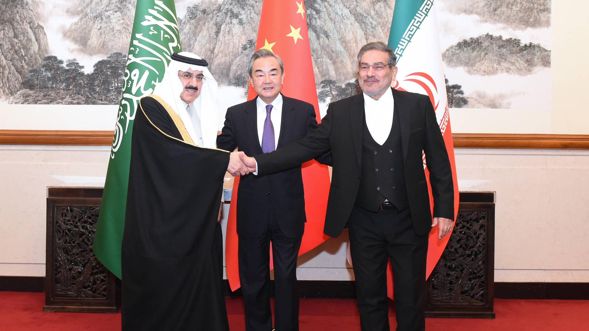 Drei Männer stehen zusammen vor den Fahnen Irans, Chinas und Saudi Arabiens. Es sind ein iranischer, ein chinesischer und ein saudi arabischer Politiker. Hinter ihnen befindet sich eine chinesische Zeichnung, das Treffen fand in China statt.