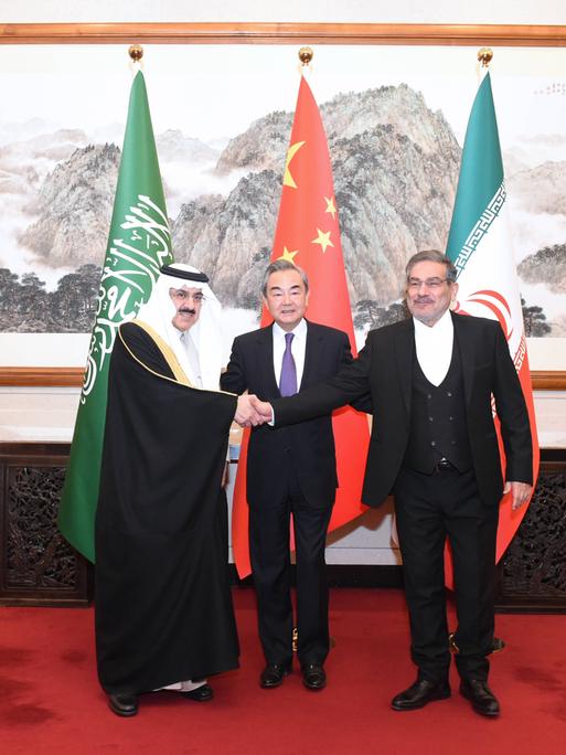 Drei Männer stehen zusammen vor den Fahnen Irans, Chinas und Saudi Arabiens. Es sind ein iranischer, ein chinesischer und ein saudi arabischer Politiker. Hinter ihnen befindet sich eine chinesische Zeichnung, das Treffen fand in China statt.