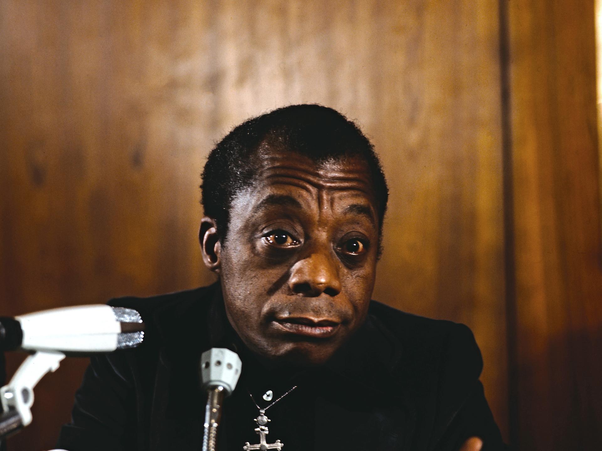 James Baldwin sitzt in schwarz gekleidet mit silbernem Kreuz um den Hals rauchend vor Mikrofonen.