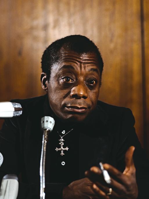 James Baldwin sitzt in schwarz gekleidet mit silbernem Kreuz um den Hals rauchend vor Mikrofonen.