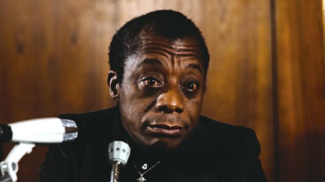 James Baldwin sitzt in schwarz gekleidet mit silbernem Kreuz um den Hals rauchend vor Mikrofonen.