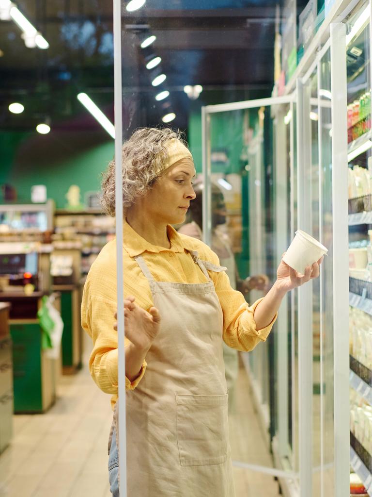 Eine Angestellte im Supermarkt hat eine Kühlvitrine geöffnet und hält einen Joghurtbecher in der Hand.