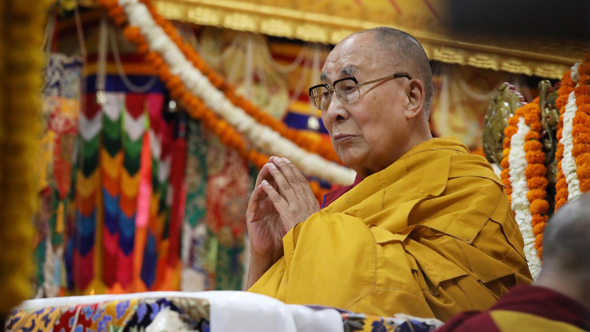 Der Dalai Lama steht in einem gelben Gewand und mit gefalteten Händen im Haupttempel in Dharamsala (Indien), im Hintergrund hängen bunte Schmuckbänder.