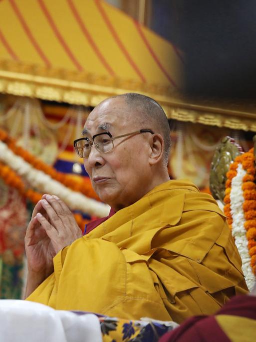 Der Dalai Lama steht in einem gelben Gewand und mit gefalteten Händen im Haupttempel in Dharamsala (Indien), im Hintergrund hängen bunte Schmuckbänder.