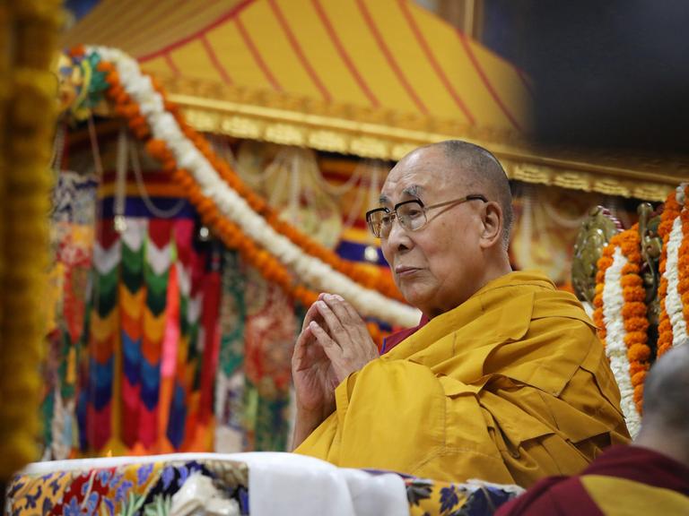 Der Dalai Lama steht in einem gelben Gewand und mit gefalteten Händen im Haupttempel in Dharamsala (Indien), im Hintergrund hängen bunte Schmuckbänder.