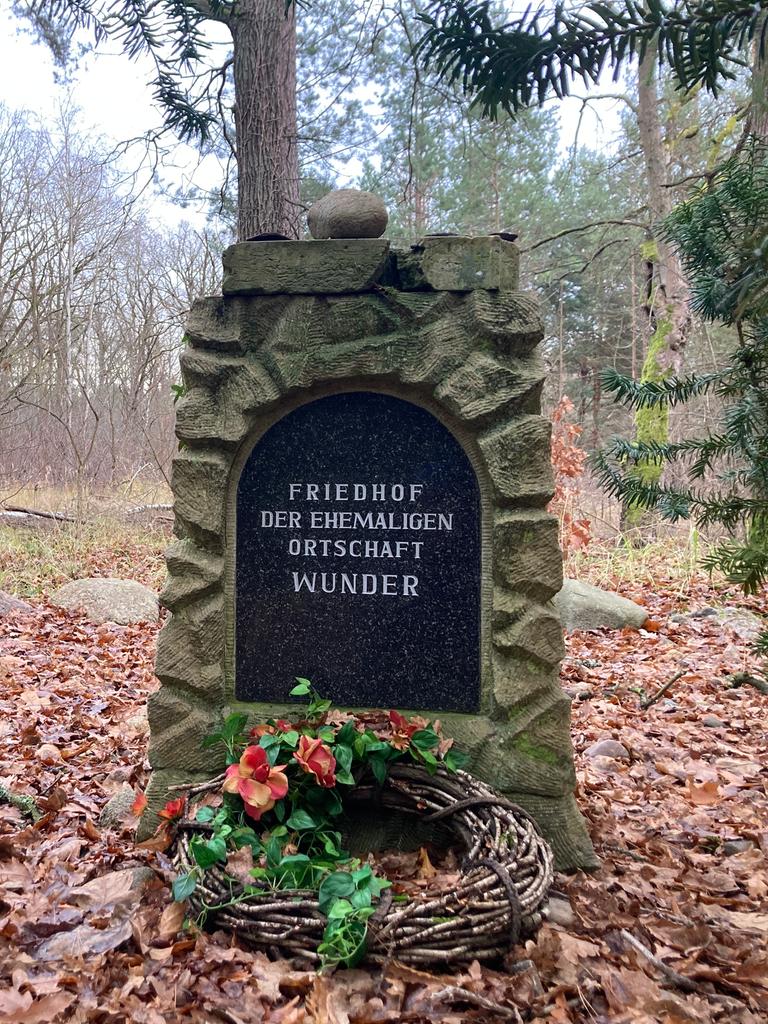 Im Wald steht ein Gedenkstein mit der Aufschrift "Friedhof der ehemaligen Ortschaft Wunder". Davor wurde ein Grabkranz abgelegt.