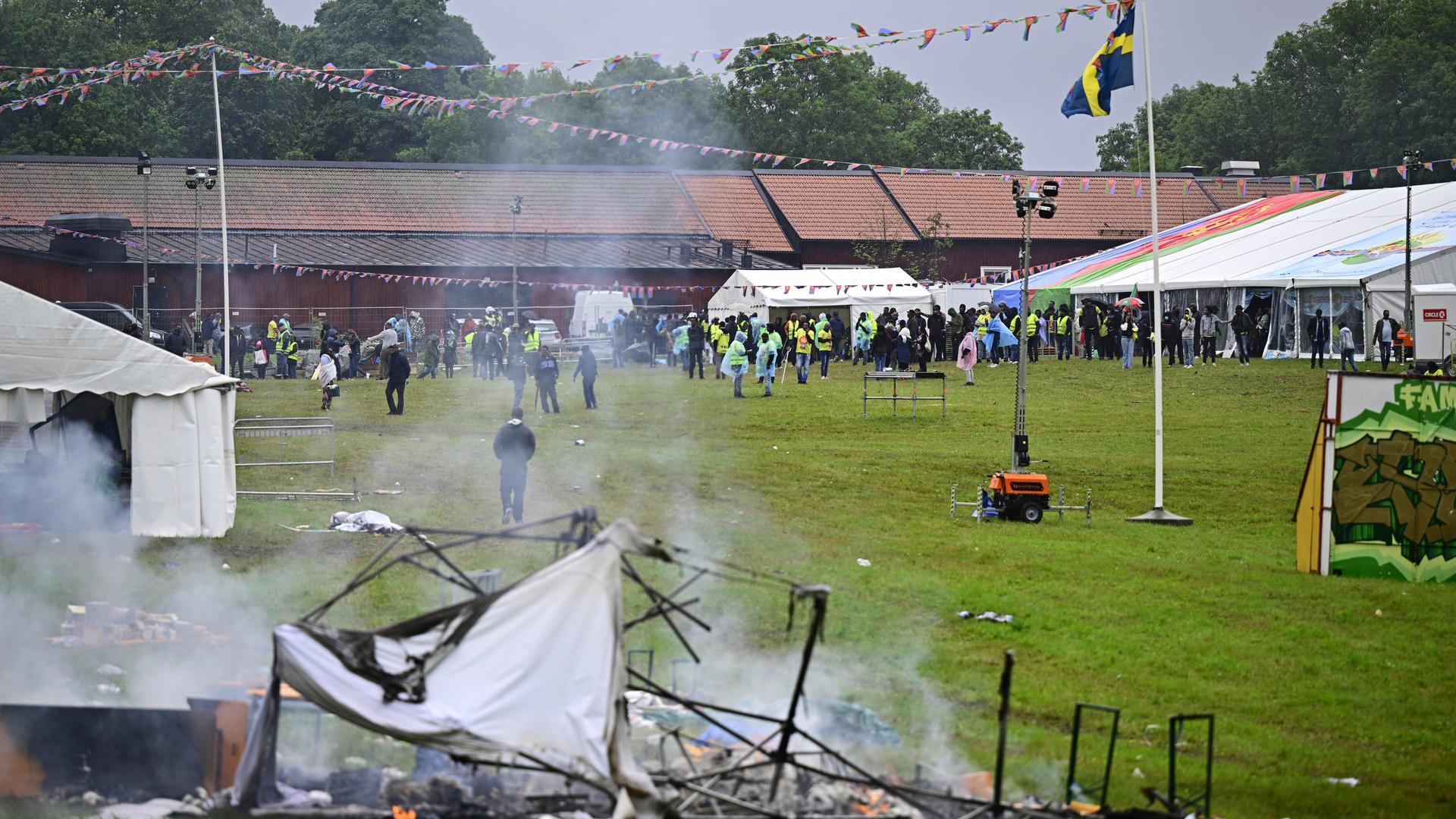 Auf einer Festwiese sind im Hintergrund Menschen zu sehen; im Vordergrund steigt von zerstörter Einrichtung Rauch auf. Eine schwedische Fahne weht an einem Mast.