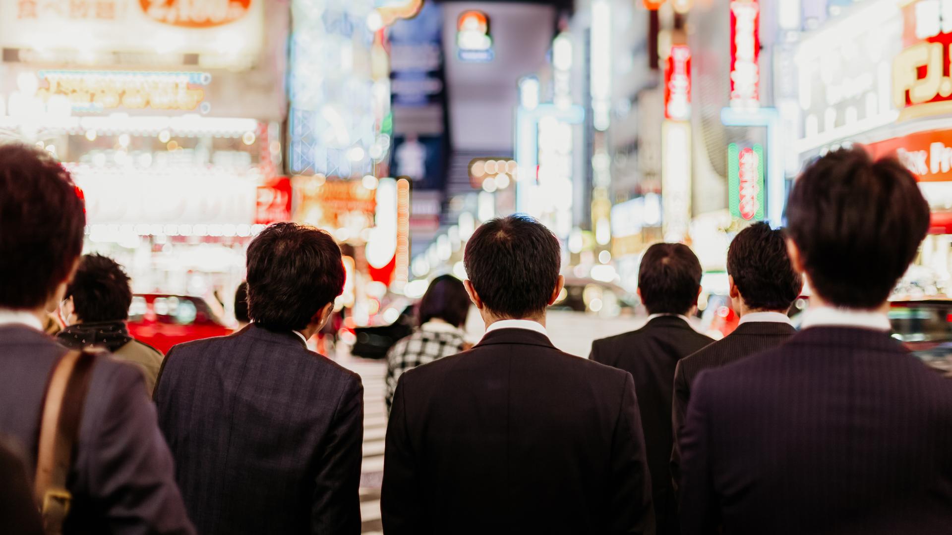 Von hinten sind japanische Geschäftsleute vor einer nächtlichen Straße mit Hochhäusern in Tokio zu sehen