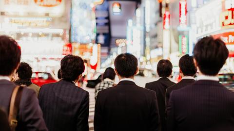 Von hinten sind japanische Geschäftsleute vor einer nächtlichen Straße mit Hochhäusern in Tokio zu sehen