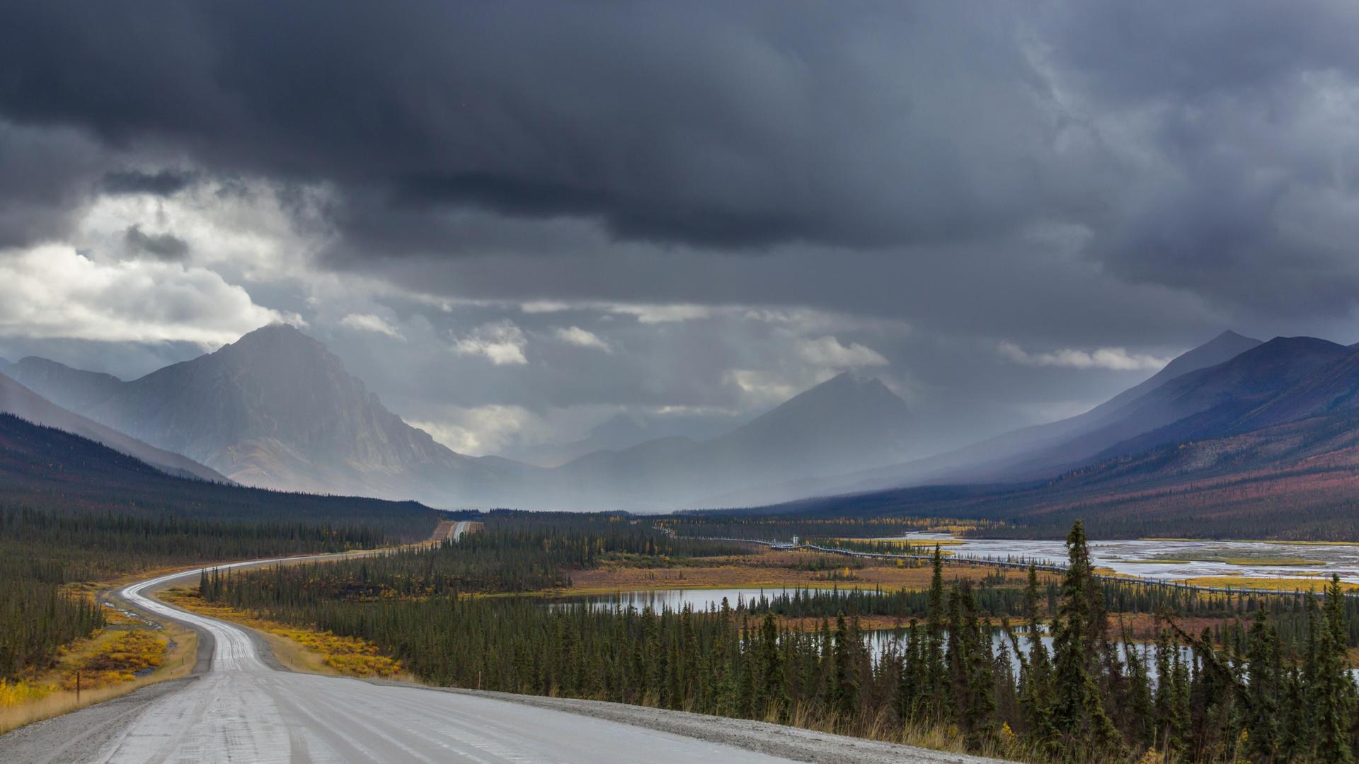 Dalton Highway in Alaska im Herbst: Eine Landstraße führt auf Berge zu, über diesen hängen dunkle Wolken.