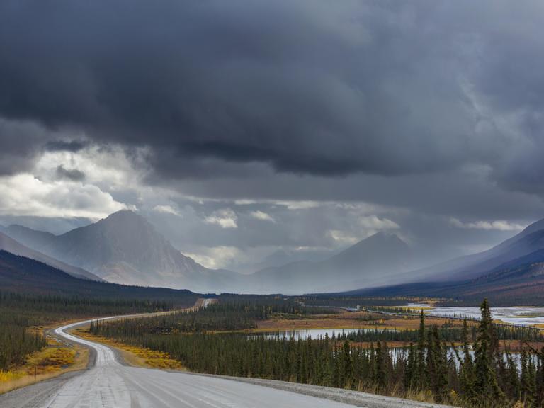 Dalton Highway in Alaska im Herbst: Eine Landstraße führt auf Berge zu, über diesen hängen dunkle Wolken.