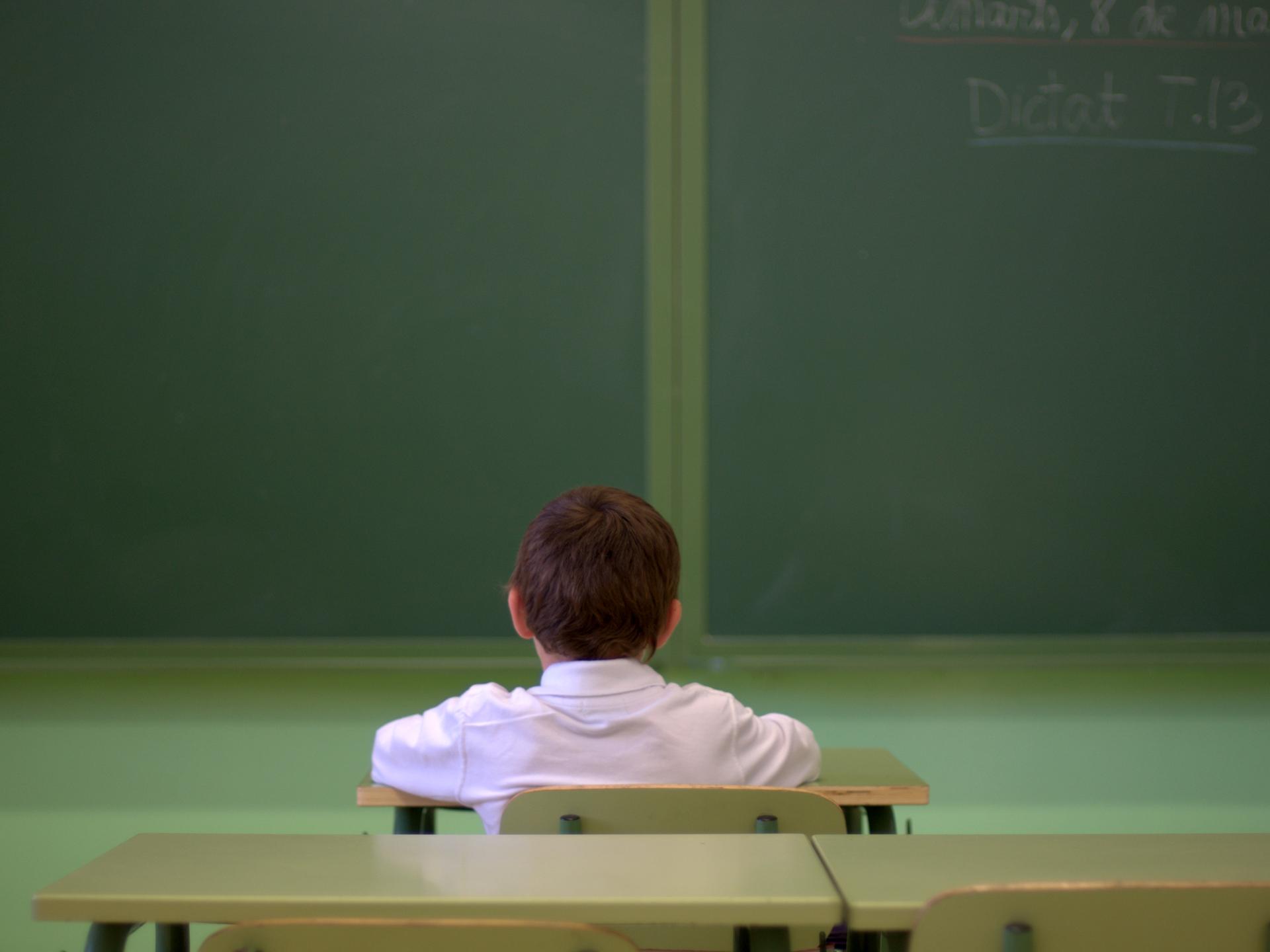 Ein Junge sitzt alleine in einem Klassenraum und guckt in Richtung der Tafel. Die Kamera hat ihn von hinten aufgenommen.