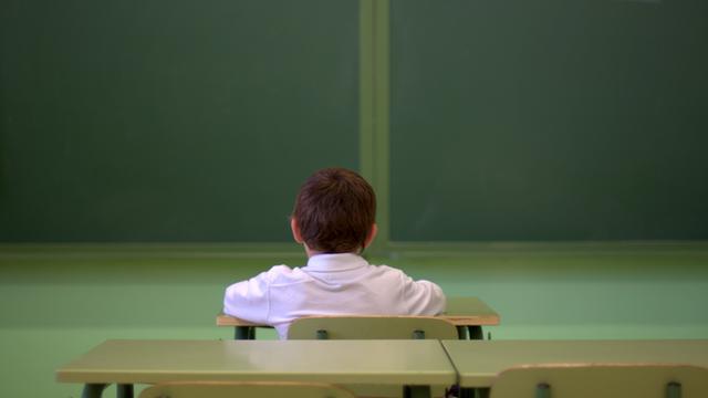Ein Junge sitzt alleine in einem Klassenraum und guckt in Richtung der Tafel. Die Kamera hat ihn von hinten aufgenommen.