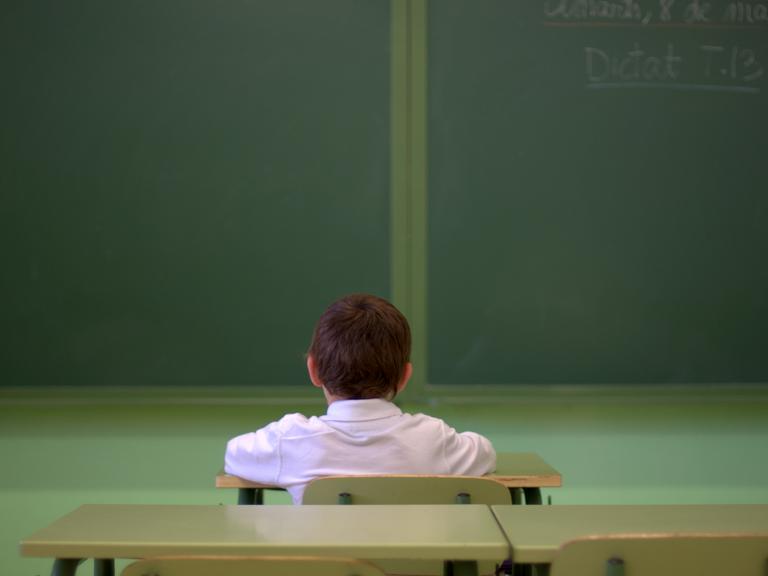 Ein Junge sitzt alleine in einem Klassenraum und guckt in Richtung der Tafel. Die Kamera hat ihn von hinten aufgenommen.