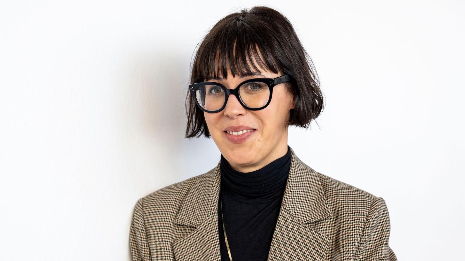 Eva Karl Faltermeier steht mit schwarzem Oberteil und kariertem Sakko vor einem weißen Hintergrund. Sie guckt durch eine schwarz umrandete Brille in die Kamera. Eva Karl Faltermeier steht mit schwarzem Oberteil und kariertem Sakko vor einem weißen Hintergrund. Sie guckt durch eine schwarz umrandete Brille in die Kamera.