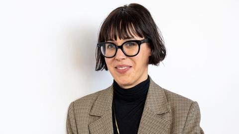Eva Karl Faltermeier steht mit schwarzem Oberteil und kariertem Sakko vor einem weißen Hintergrund. Sie guckt durch eine schwarz umrandete Brille in die Kamera.